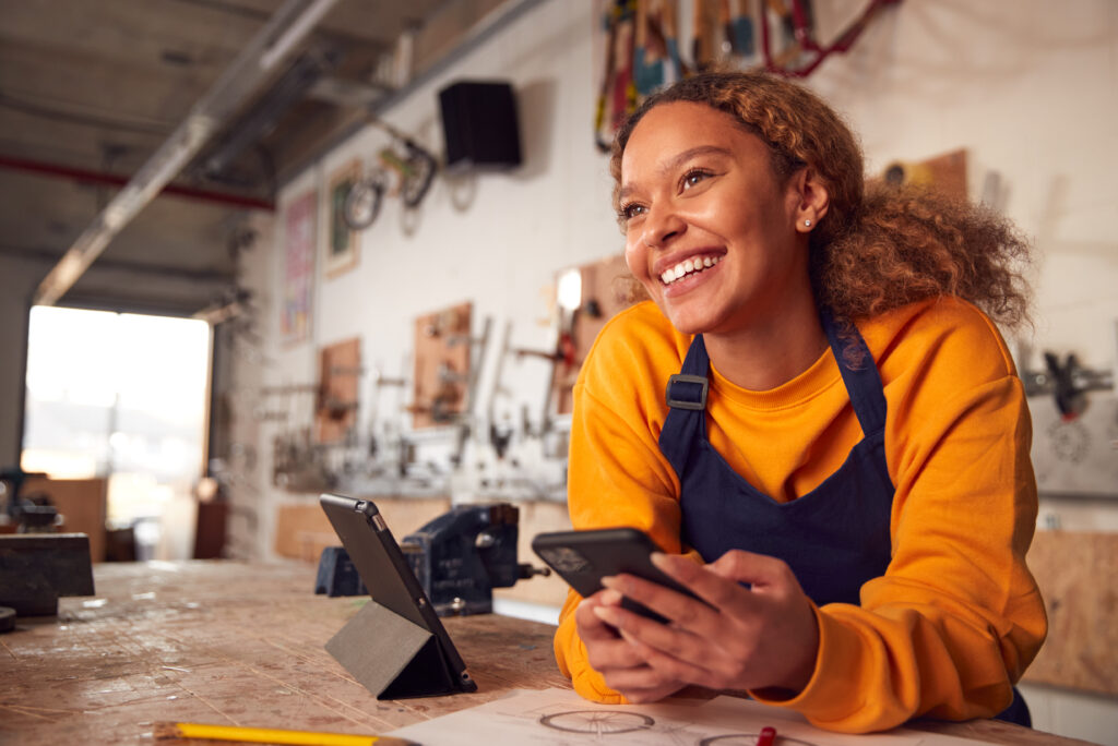 Female Business Owner In Workshop Using Digital Tablet And Holding Mobile Phone