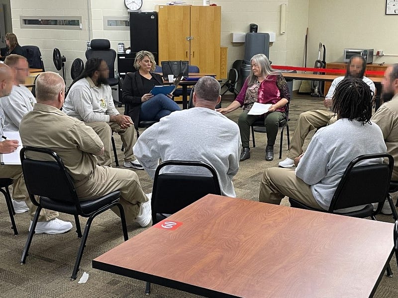 A group of diverse individuals sit in a circle listening to two women speak. Most of the individuals wear a combination of gray sweatshirts and khaki pants.