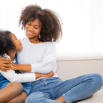 African American young mother embrass her daughter with smile of love, looking each other on sofa at home, Black couple woman and kid express feelling of love.