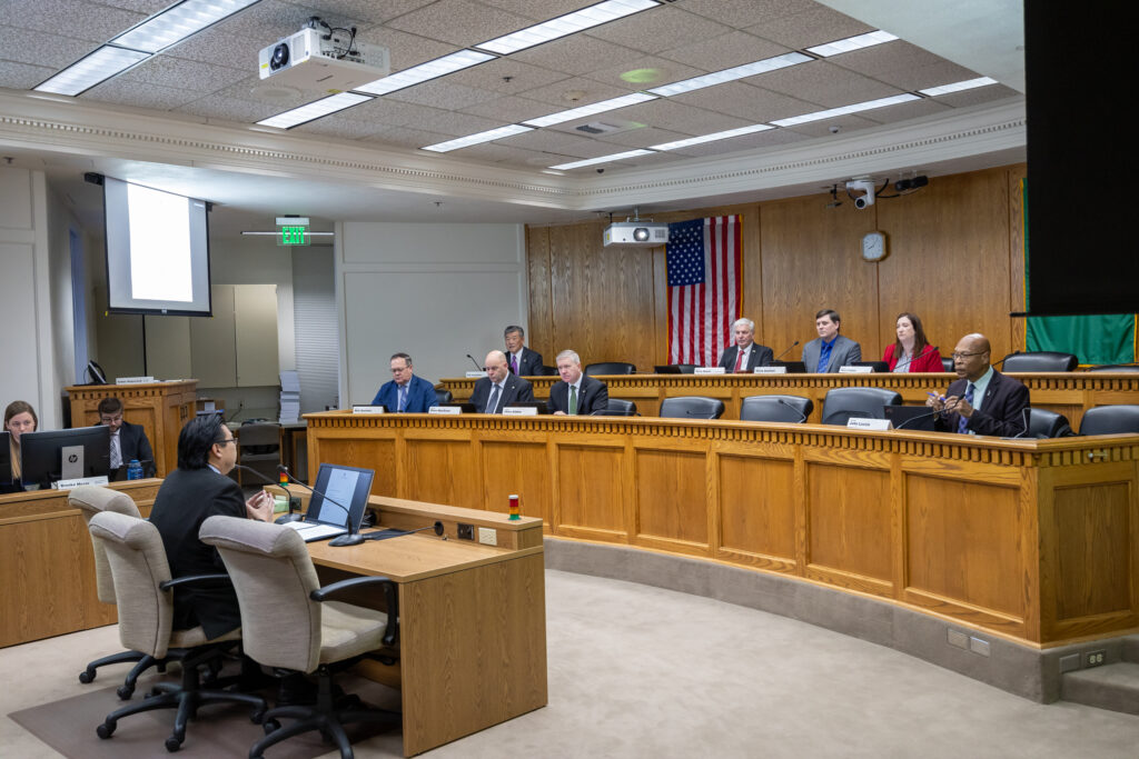A legislative meeting room with several officials seated at a two-tiered bench. A person sits behind a desk and speaks in front of them. Two other people to the side are seated behind computer screens. An American and Washington state flag are visible on the wall behind the panel of lawmakers. 