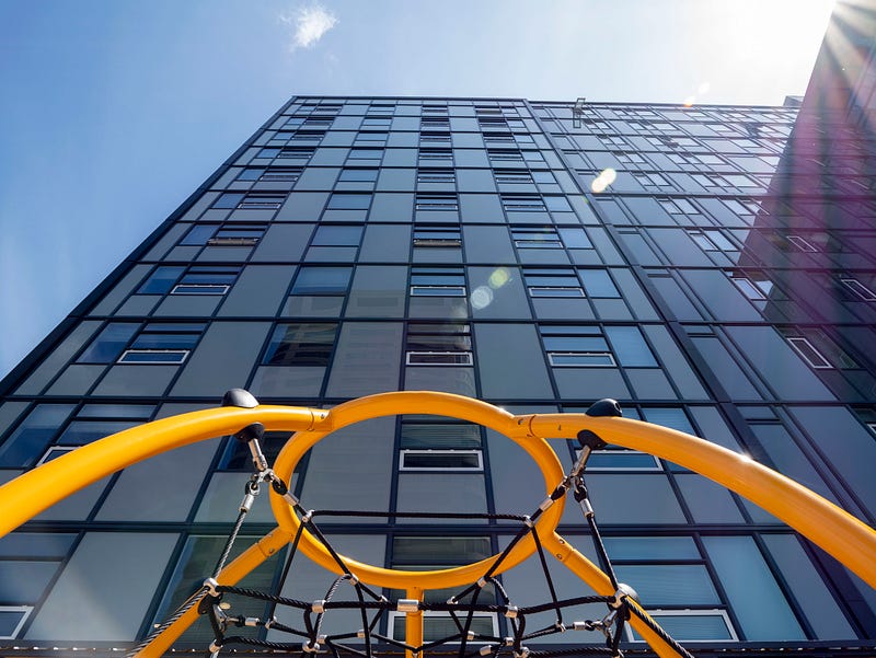 A yellow play structure is seen in the foreground in front of a towering high-rise apartment building. Blue sky with a bright sunburst are behind the building. 