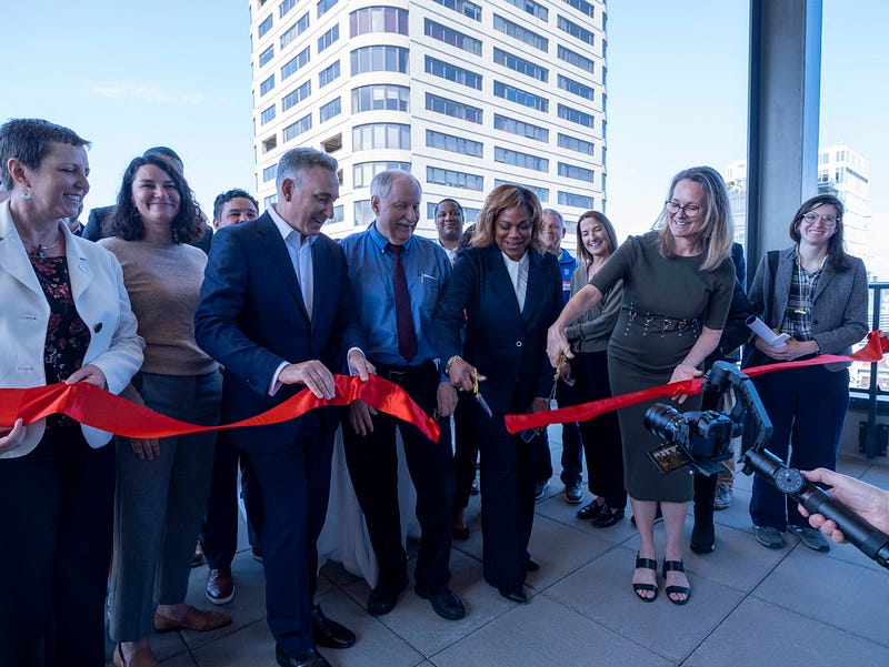 A group of people stand holding a red ribbon as it is cut by two women holding scissors in the foreground. A high-rise building is visible in the background. 
