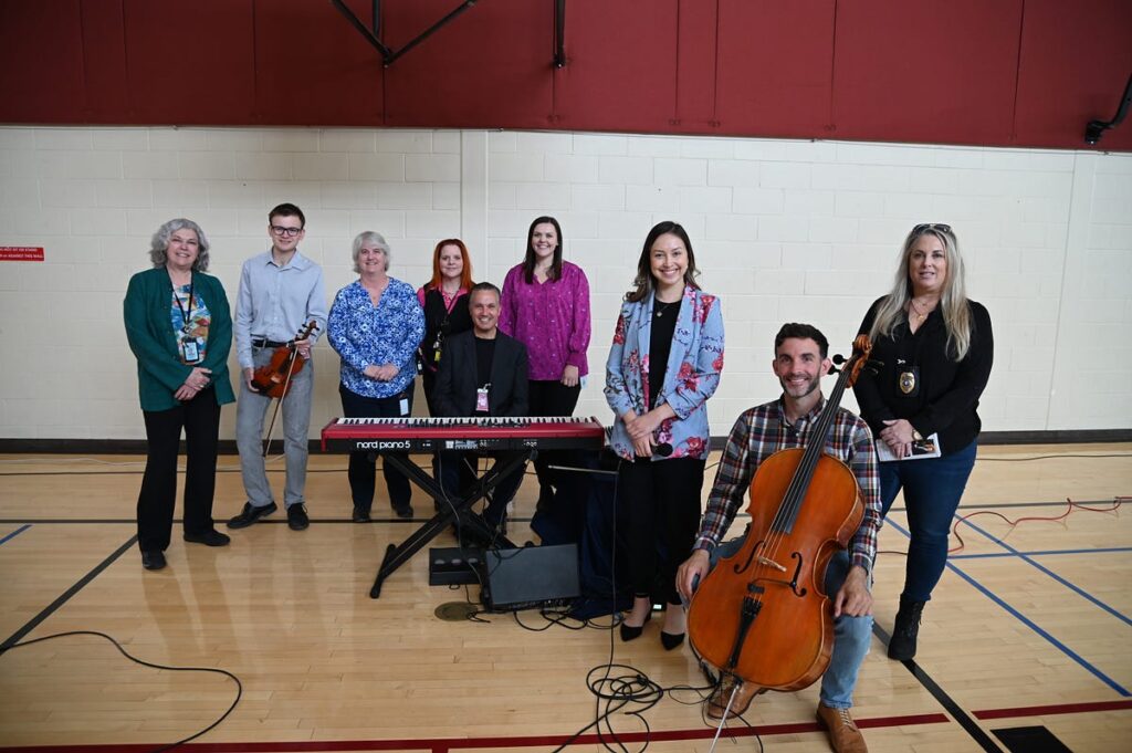 Nine individuals pose together in a gymnasium for a concert. One sits in the foreground with a cello, another is in the center behind keyboards.