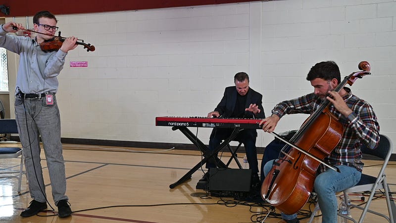 Three musicians perform together in a gymnasium setting. Each play a different instrument: Violin, keyboards and cello. The violin player on the left is the only person standing.