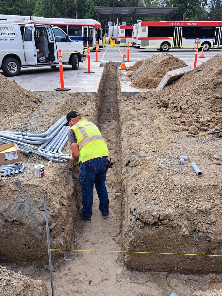 A worker in a safety vest and wearing a ball cap digs a trench for EV charging infrastructure. There are pipes laying next to the trench and Jefferson Transit vehicles in the background, as well as a white van with the side doors open.
