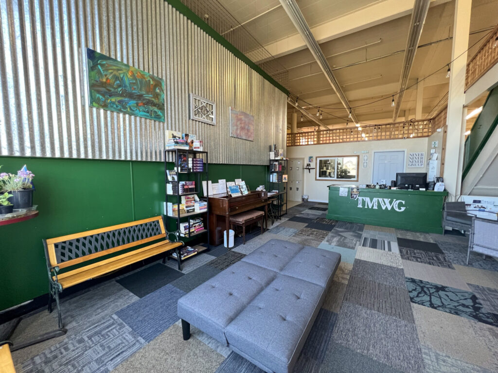 The interior of The Moore Wright Group's Recovery Café of Hope. The lower walls and welcome counter are dark green with a collection of benches and bookshelves throughout. Paintings by TMWG staff hang on the walls.