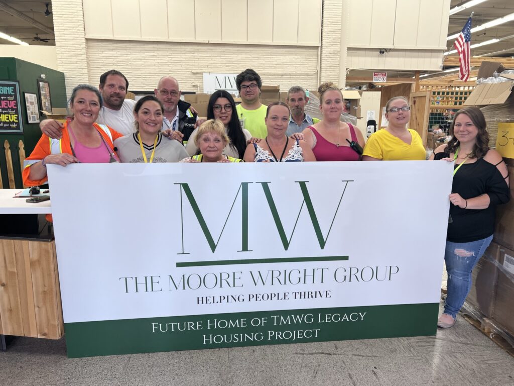 A dozen staff members from The Moore Wright Group pose together in the organization's warehouse behind a white sign with green font announcing their Legacy Housing.