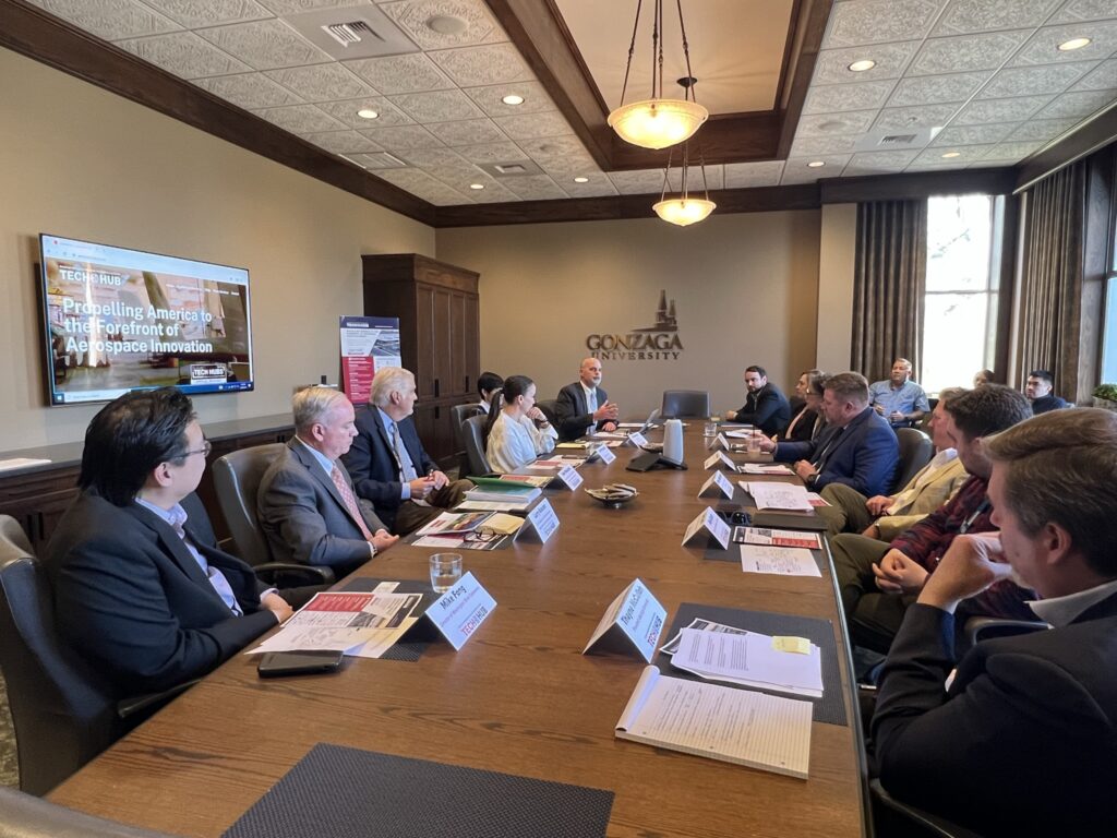 A group of people sit around a boardroom table at Gonzaga University. Three people sit in the right back corner against the window. A TV screen displays a slideshow to the left.