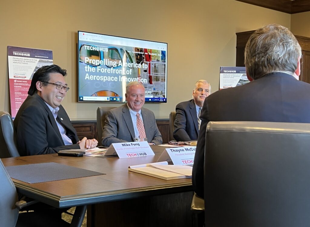 Four people at a boardroom table with a slideshow playing on a screen behind them. Two of them are laughing.