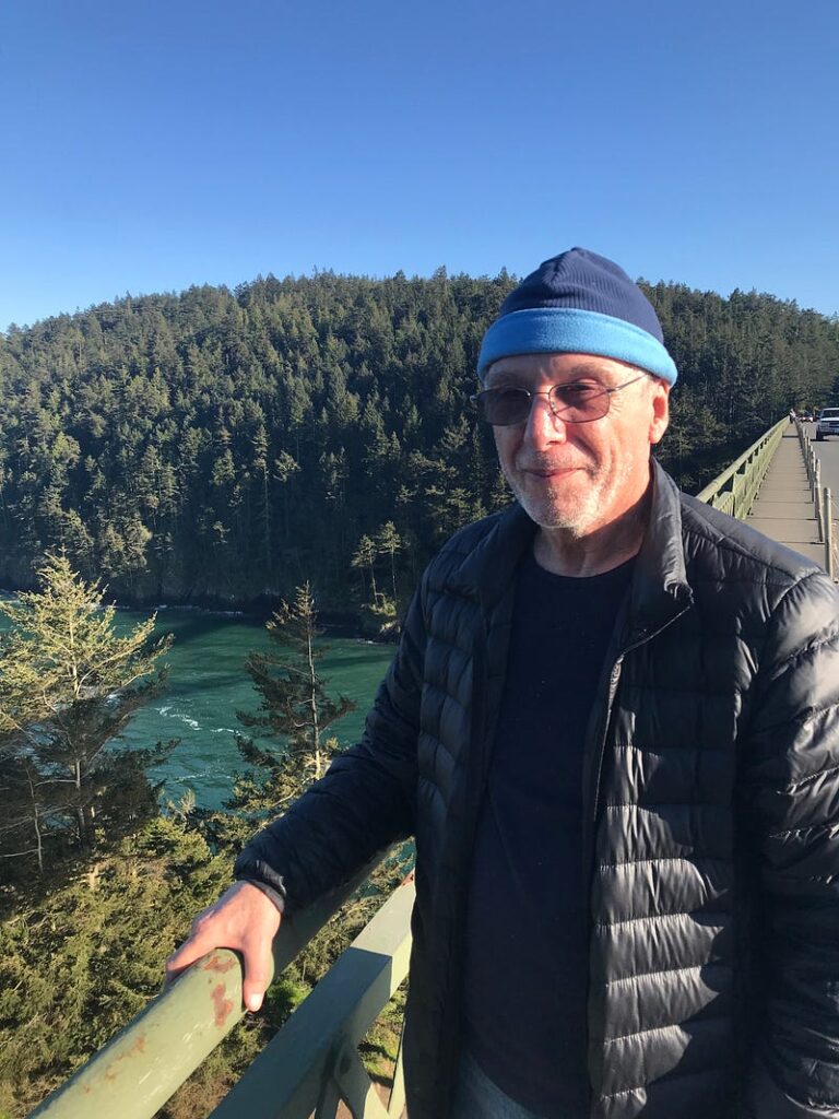 A man wearing a coat and beanie cap stands on a bridge overlooking water and forested area.