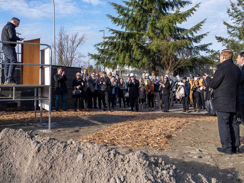 A group of people gather in front of trees at a construction site. They listen to a speaker on a makeshift podium on the left side. 