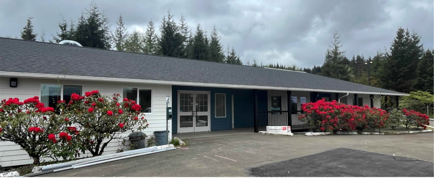 The exterior of Alder House, a single-story building with red flowers framing the entrance and tall trees in the background under a cloudy sky.