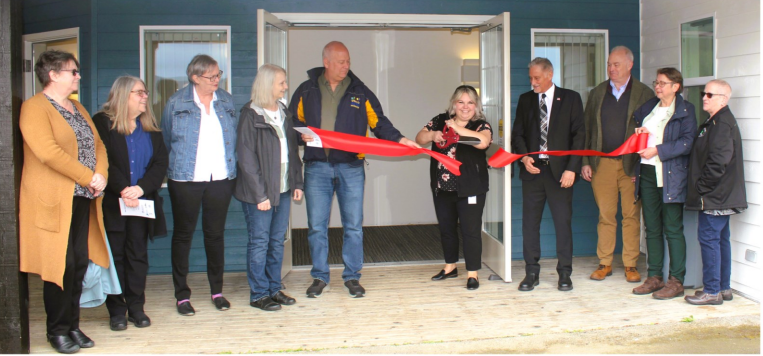 A group of housing advocates stands in front of the door to Alder House, with one person cutting a red ribbon.