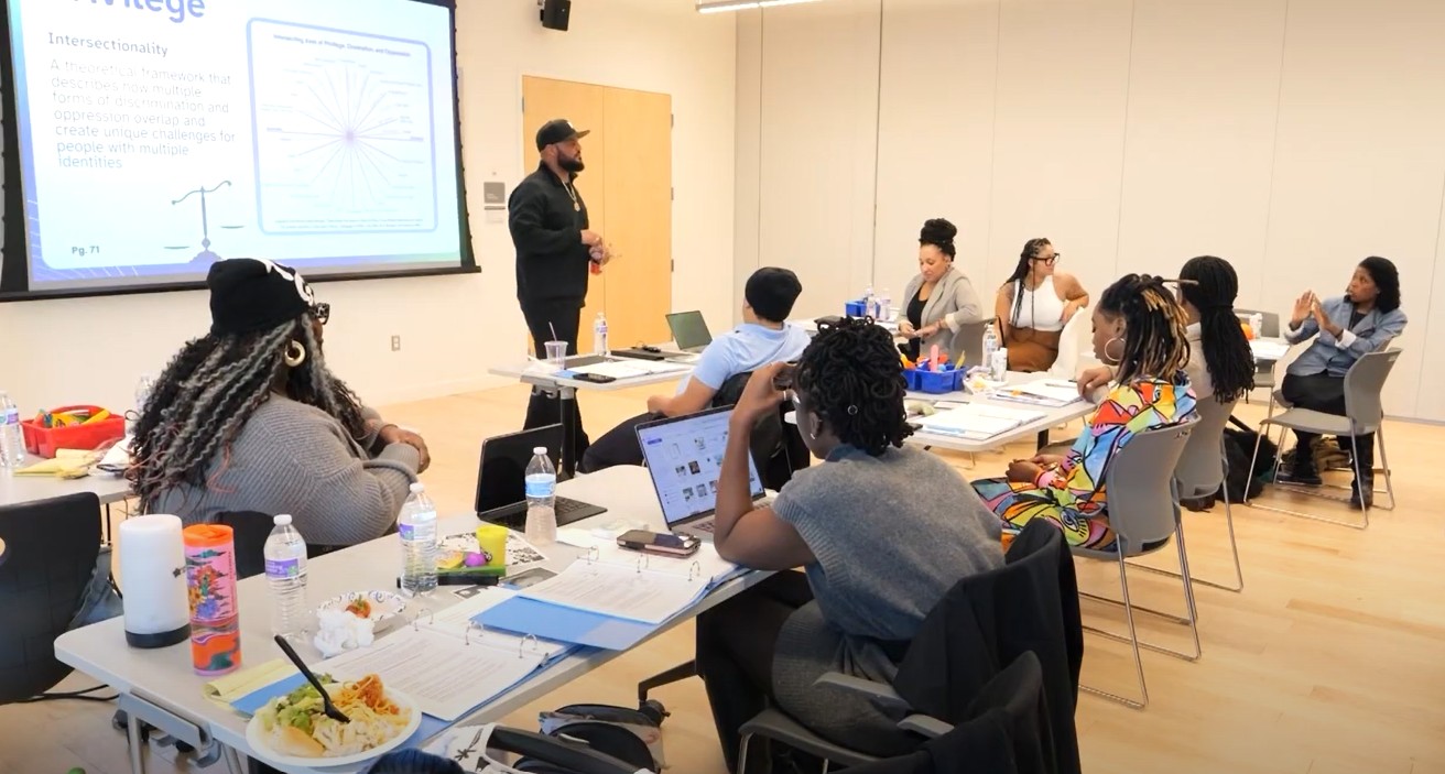 People in a beige room learning about supporting their peers. They are sitting around a table while a gentleman speaks to them and leads the conversation.